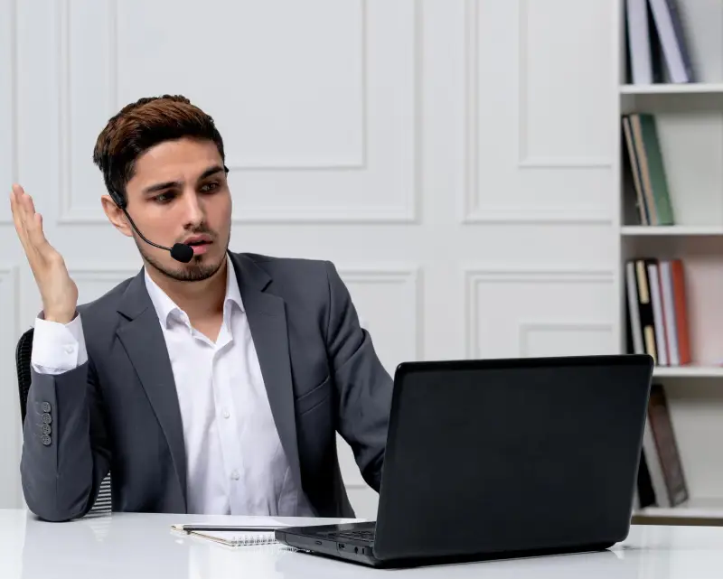A business professional in a suit gestures while sitting at a desk with a laptop, surrounded by bookshelves and decorative items